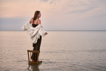 Young woman stands gracefully on chair in calm water, framed by serene sunset. Her pose evokes...