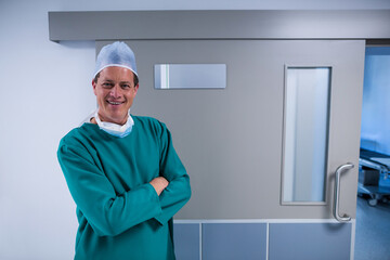 Man in green scrubs, surgical cap, mask standing in hospital hall by stretcher door, copy space