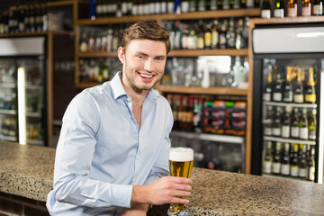 Man sitting at bar counter holding glass of beer, smiling near glass-door cooler and wooden shelves