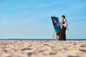 Young female painter works on canvas at serene beach. Tranquil setting of shoreline and open sea...