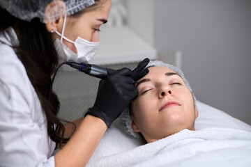 Young woman receives eyebrow permanent makeup in serene spa. Beauty technician, wearing gloves,...
