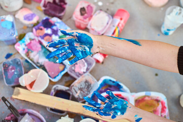 Adult hands covered in vivid blue paint, with scattered art supplies outdoors. Soft lighting casts...
