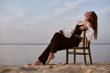 Young female artist sits on seaside beach, leaning back in chair with paintbrush in hand. Scene...