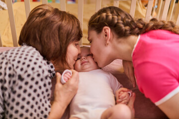 Elderly woman with brown hair and young woman with braided hair, both affectionate, lean toward infant inside crib. Soft lighting, warm, intimate atmosphere