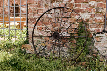 Old rusty metal wagon wheel resting against weathered brick wall, surrounded by overgrown grass and...