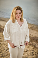 Young woman in casual white attire stands on tranquil beach, exuding calm as waves gently lap...
