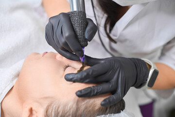 Professional applies eyebrow permanent makeup to young woman in clinic setting. Use of specialized...