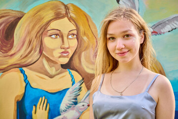Young blonde female standing confidently before colorful mural. Artwork depicts woman, bird, blue dress. Sunny day, soft shadows enhance details beautifully