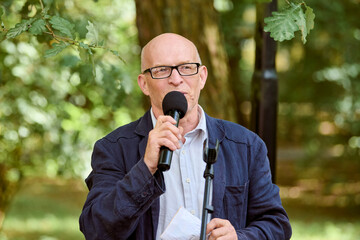 Bald mature man with glasses delivers outdoor lecture about amber jewellery. Man holds microphone,...