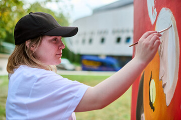 Young caucasian woman in black cap painting vibrant mural outdoors. Bright daylight and green...