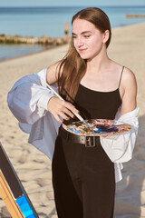 Young female painter stands on tranquil beach, embracing calming ambiance as she holds palette. Her...
