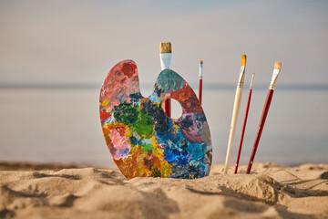 Vibrant painter palette and brushes rest on tranquil beach, symbolizing creativity and inspiration....