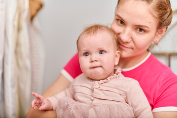 Young mother with light brown hair, holding baby girl. Woman wears pink top, baby in light dress. Warm emotions, indoor environment, bright lighting