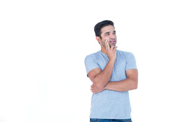 Man standing on plain white backdrop wearing grey t-shirt, blue jeans, holding cheek thoughtfully