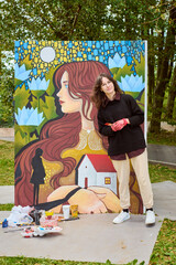 Young female with wavy brown hair standing by colorful en plein air canvas. Smiling, holding...
