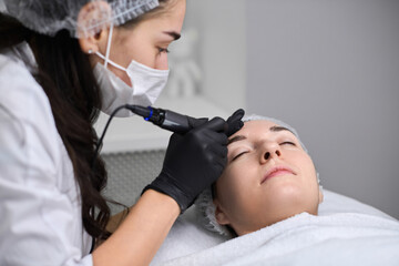 Young woman receives eyebrow permanent makeup in serene spa. Beauty technician, wearing gloves,...