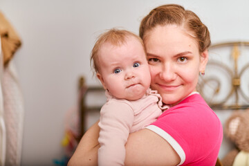 Smiling young mother with light brown hair holds baby girl. Indoor setting with soft lighting. Background with warm tones. Baby curious expression