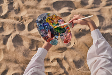 Young adult female artist uses paintbrush on colorful palette while seated on sunlit sandy beach....