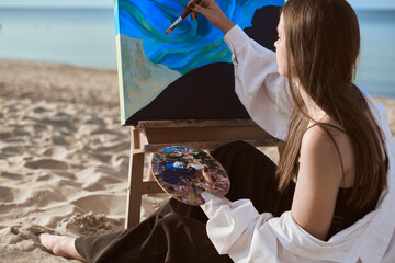 Young woman artist paints by sea, blending colors on palette. Her serene expression reflects...