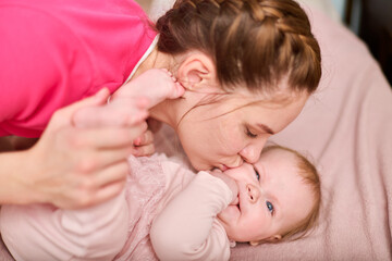 Young woman with brown hair kisses infant daughter with light hair on bed. Warm, gentle moment captured in cozy setting, pink tones prominent