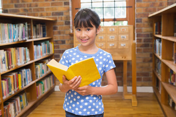 Asian girl standing in library aisle reading yellow book near card catalog by window
