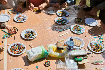 Sunlit workshop scene featuring multiple paper plates with vibrant mosaic tiles. Hands actively arrange pieces on light wooden table, creating dynamic patterns