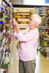 Senior female shopper reaching for jar on supermarket aisle shelves while pushing cart with produce