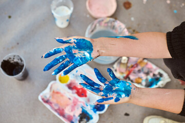 Adult hands covered in vivid blue paint, with scattered art supplies outdoors. Soft lighting casts gentle shadows on ground, enhancing artistic mood