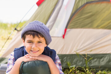 Middle Eastern boy leaning on rolled-up sleeping bag at grassy campsite beside beige green tent