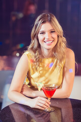 Woman sipping red cocktail from martini glass with umbrella garnish at table in dimly lit bar