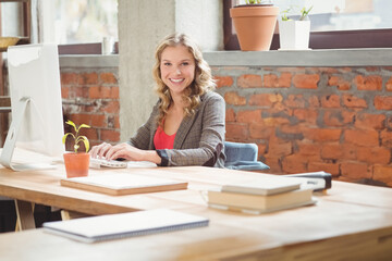 Teenage girl smiling while working at loft-style office desk with large monitor and potted plant