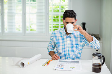 Middle Eastern man sitting at kitchen countertop drinking coffee from mug and reviewing layout