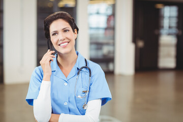 Nurse speaking on smartphone in hospital corridor, wearing light blue scrubs with stethoscope