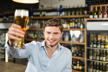 Man in twenties raising pilsner glass of beer in toast at bar counter with liquor shelves