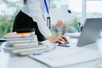 Young female employee analyzing documents and working on laptop, sitting at desk in modern office.