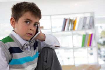 School-age boy holding smartphone to ear and gazing upward in study by shelving unit, copy space