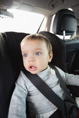 Infant boy sitting in car seat secured by black five-point harness beside back seat car headrest