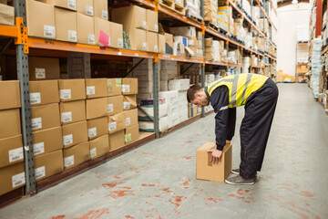 Male worker in high-visibility vest bending and lifting cardboard box on aisle with shelving racks