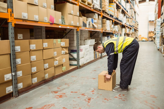 Male worker in high-visibility vest bending and lifting cardboard box on aisle with shelving racks