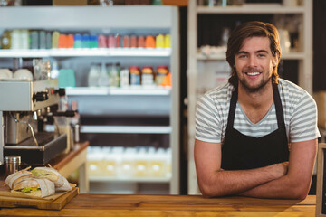 Male barista leaning on counter and smiling in coffee shop by wrapped sandwiches, espresso machine