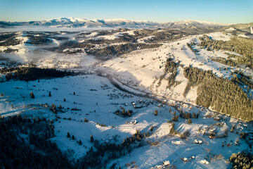 Aerial view of snow-covered valley with winding road passing through small village. Landscape features gently rolling hills, dense forests, and distant mountains under clear blue sky, with soft mist.