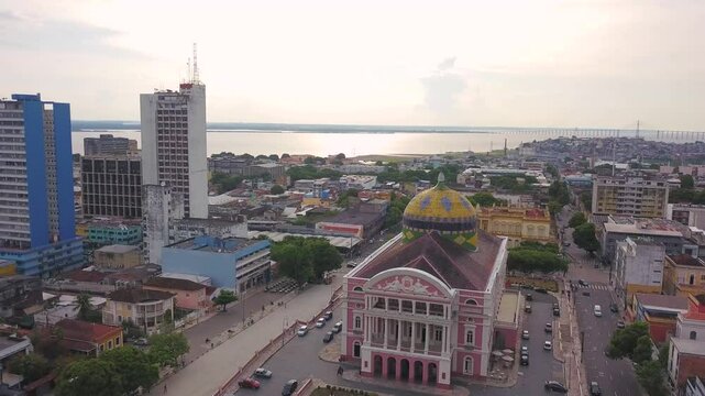 Teatro Amazonas em destaque com sua c&uacute;pula colorida, cercado pela cidade de Manaus e com o imponente rio Negro ao fundo, unindo cultura e natureza amaz&ocirc;nica.