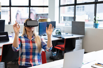 Office worker wearing VR headset raising hands at open plan office desk with laptop and monitors