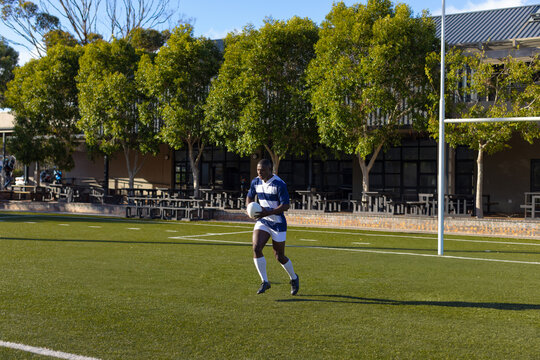 African American rugby player in jersey running across turf field holding rugby ball by goal post