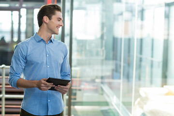 Fototapeta premium Man wearing blue shirt standing in office corridor holding tablet and smiling to left, copy space