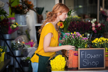 Female florist arranging yellow mums, daisies on wooden counter with chalkboard sign in flower shop