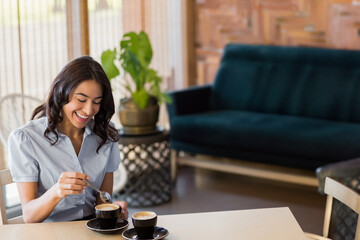 Woman in late twenties stirring coffee at café lounge table with black cups and potted plant