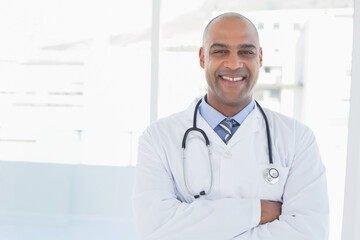 Male doctor standing in clinic corridor with floor-to-ceiling windows, wearing stethoscope and coat