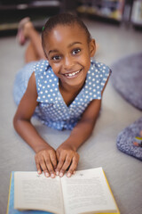 African American child girl lying on floor in living area reading book beside shelves and rug