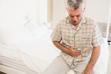 Man wearing plaid pajamas sitting on bed and clutching abdomen in bright bedroom with lamp